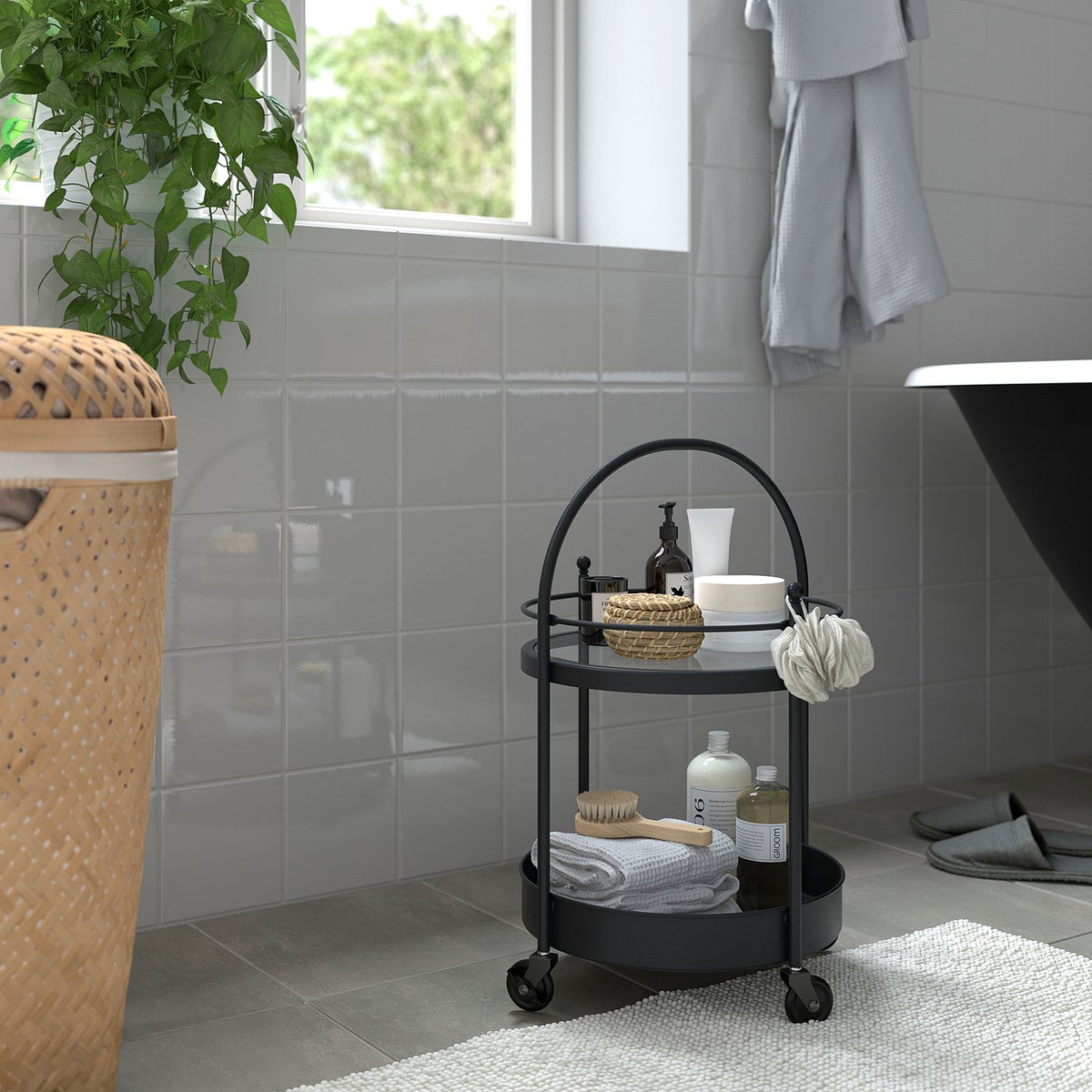 Black FRÖSJÖN bathroom trolley with towels, bottles, and a brush by bathtub and window.