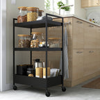 Black NISSAFORS kitchen trolley with three shelves, holding food in jars and utensils, fitting in small spaces with four wheels.