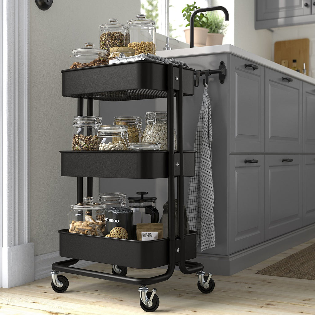 Black three-tiered RÅSKOG trolley in kitchen, holding jars and food, on wheels, compact size.