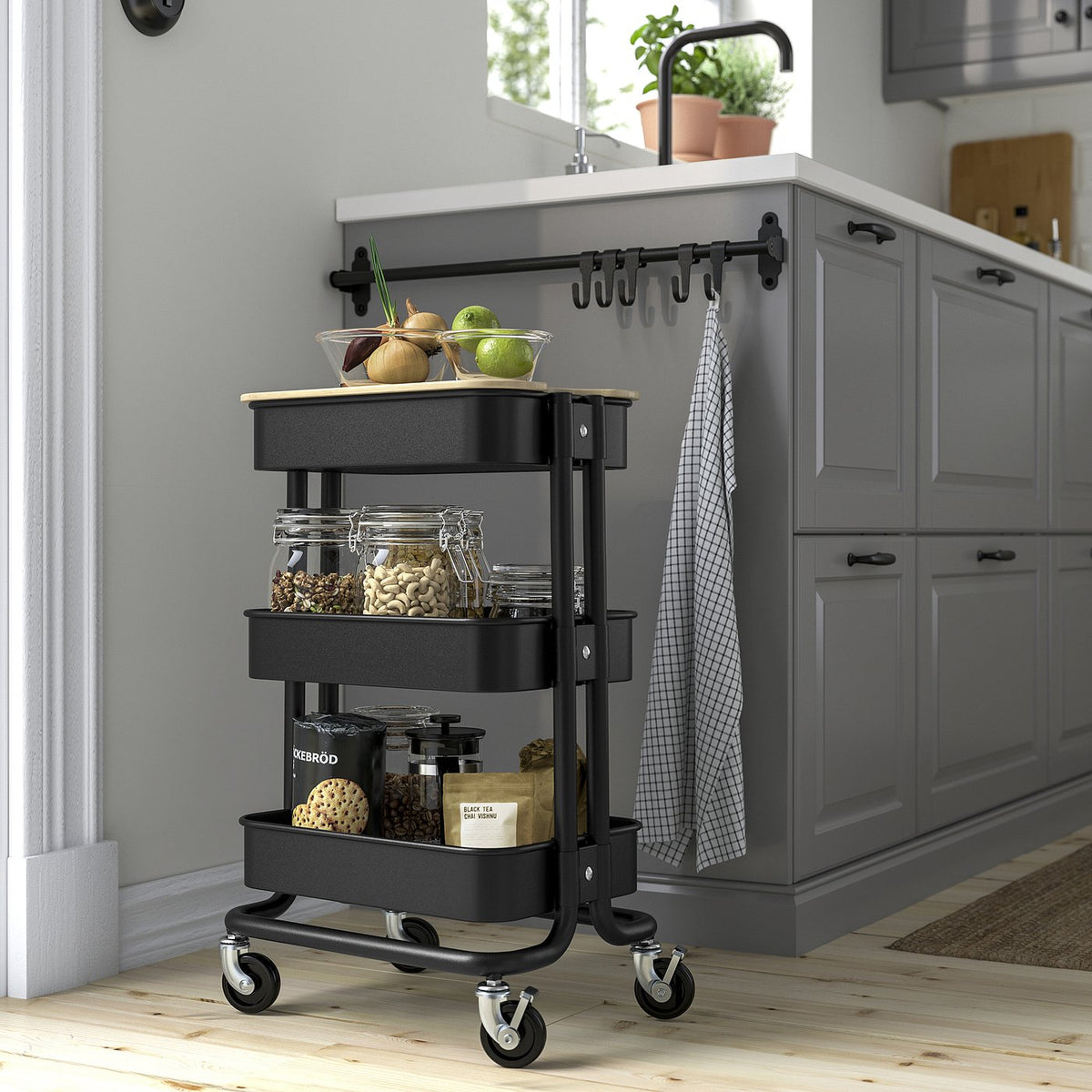 Black RÅSKOG trolley with 3 shelves, various kitchen items, grey kitchen cabinets, and a window with a plant, suitable for small spaces.